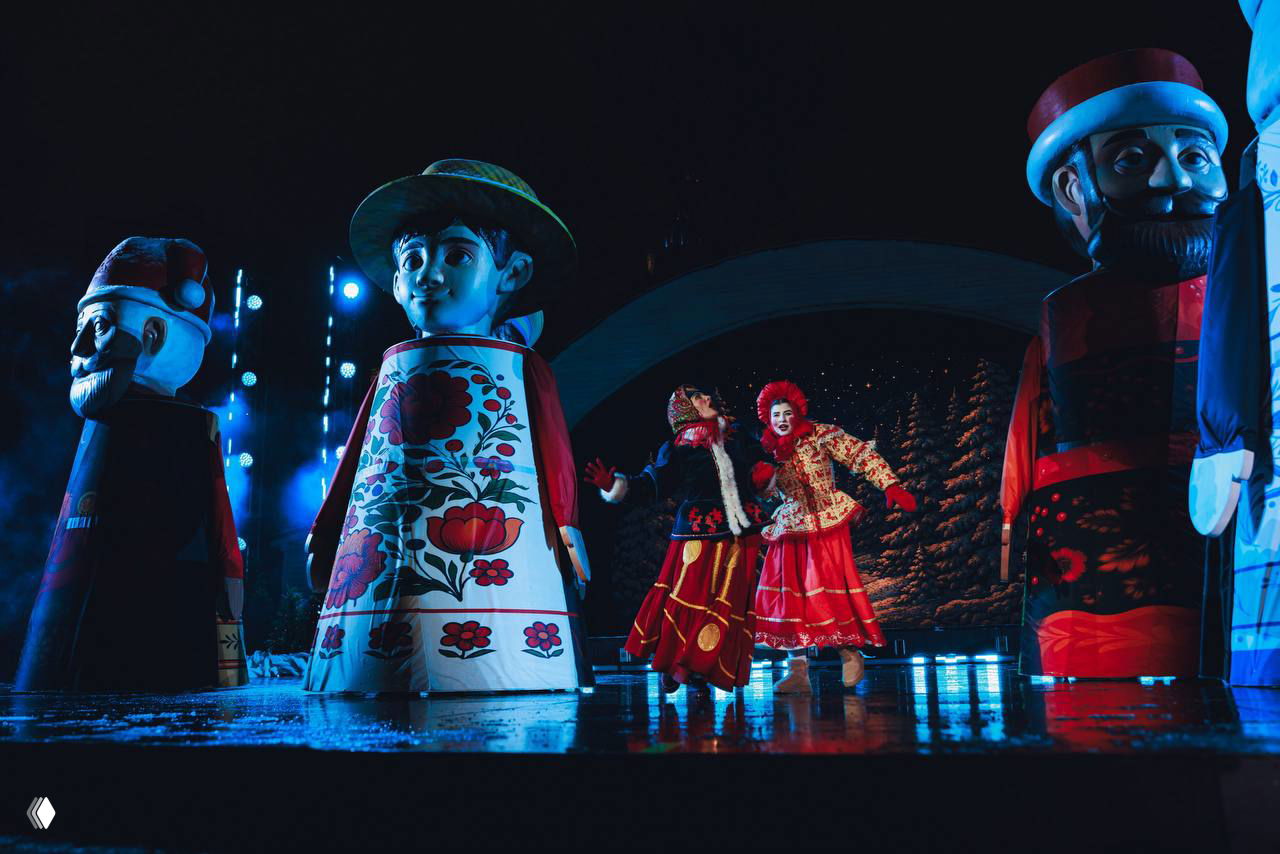 Артисты в ярких народных костюмах на фоне больших фигур в русском стиле — сцена музыкального спектакля на открытой площадке.
