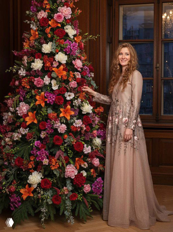 Женщина в длинном платье стоит рядом с высокой композицией из живых ярких цветов в уютном деревянном интерьере, улыбается.