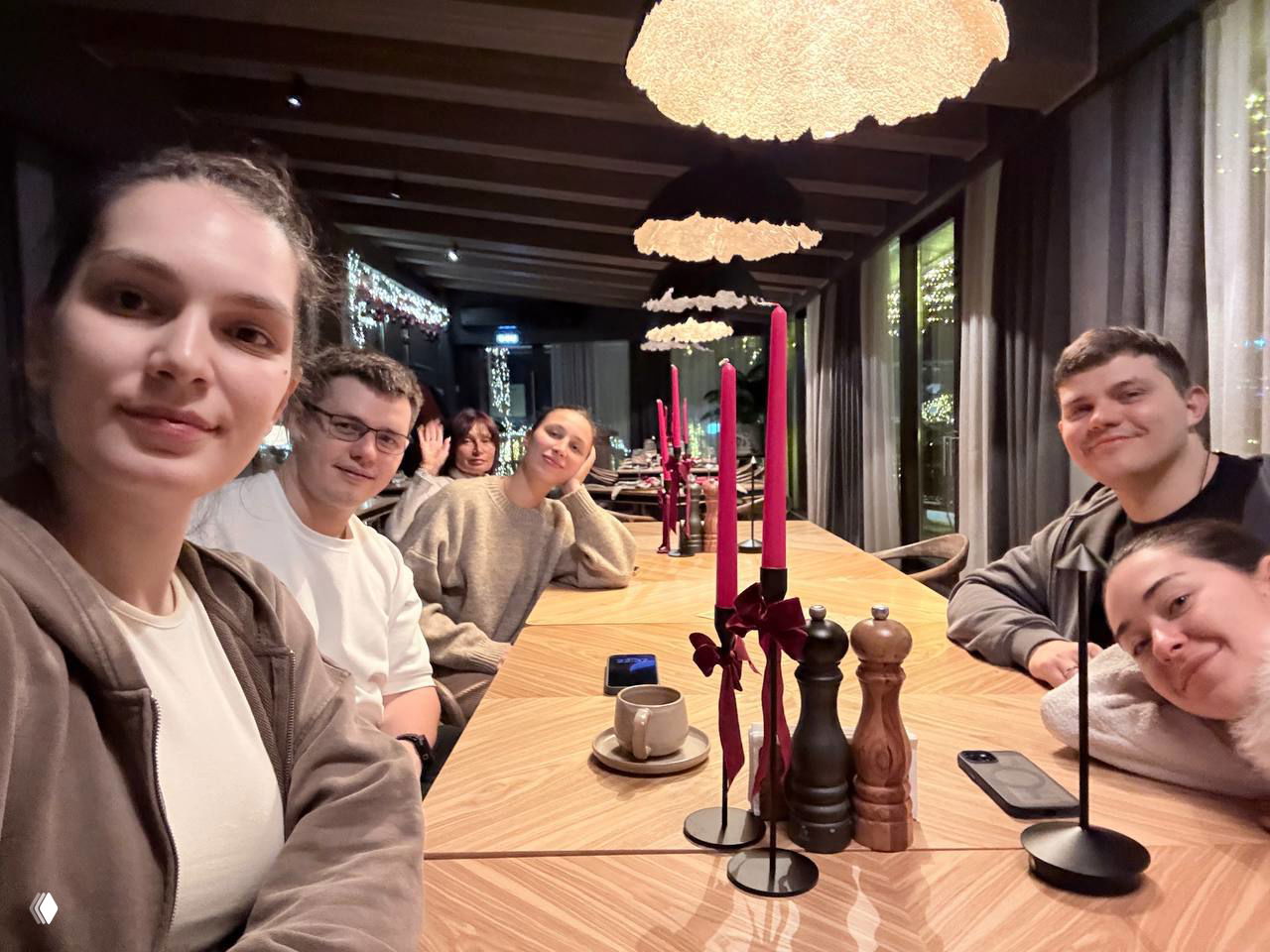 Групповое фото за длинным столом в ресторане: несколько друзей улыбаются, на столе свечи и посуда, тёплая атмосфера ужина во время отпуска.