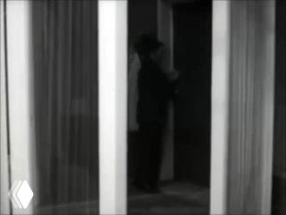 Черно-белая фотография: силуэт человека стоит в дверном проёме за стеклянными или жалюзийными перегородками, создавая атмосферу изоляции и задумчивости.