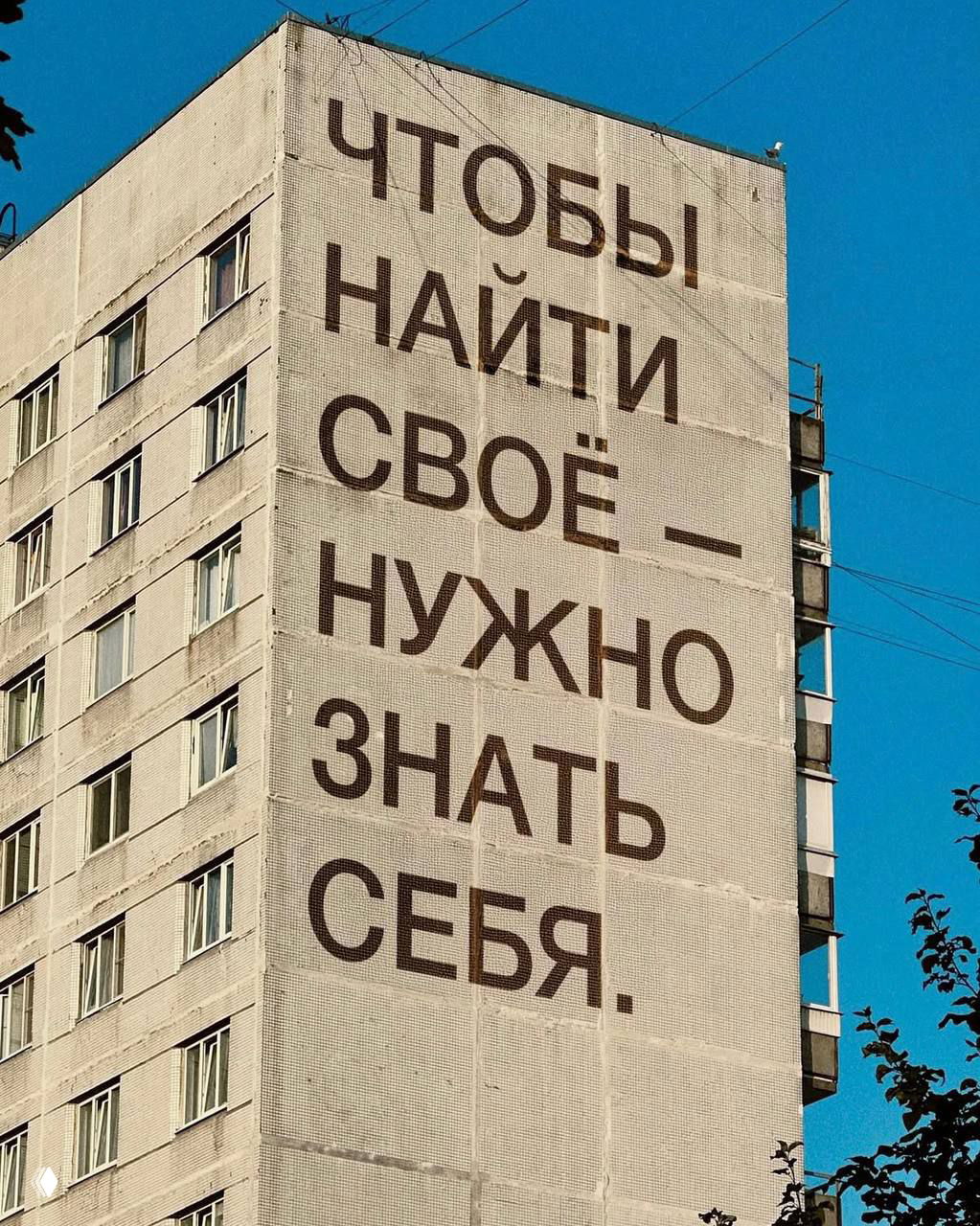 Фасад многоэтажного дома с крупной мотивирующей надписью на русском языке на фоне голубого неба; урбанистическая фотография с текстом о поиске себя.