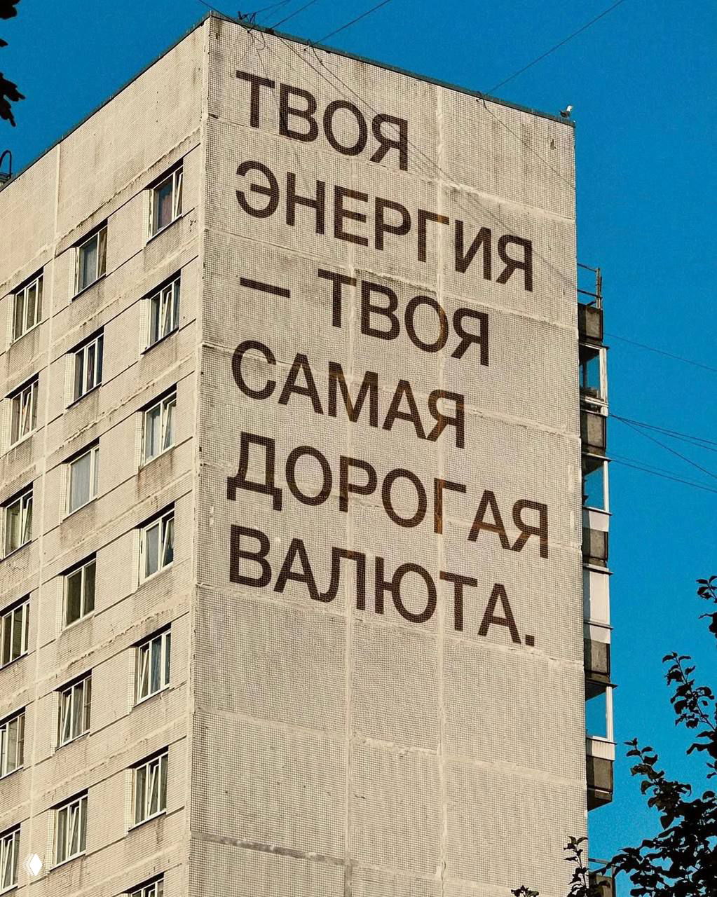 Большая надпись на стене жилого дома: «Твоя энергия — твоя самая дорогая валюта», фотография с насыщенным голубым небом и городским пейзажем.