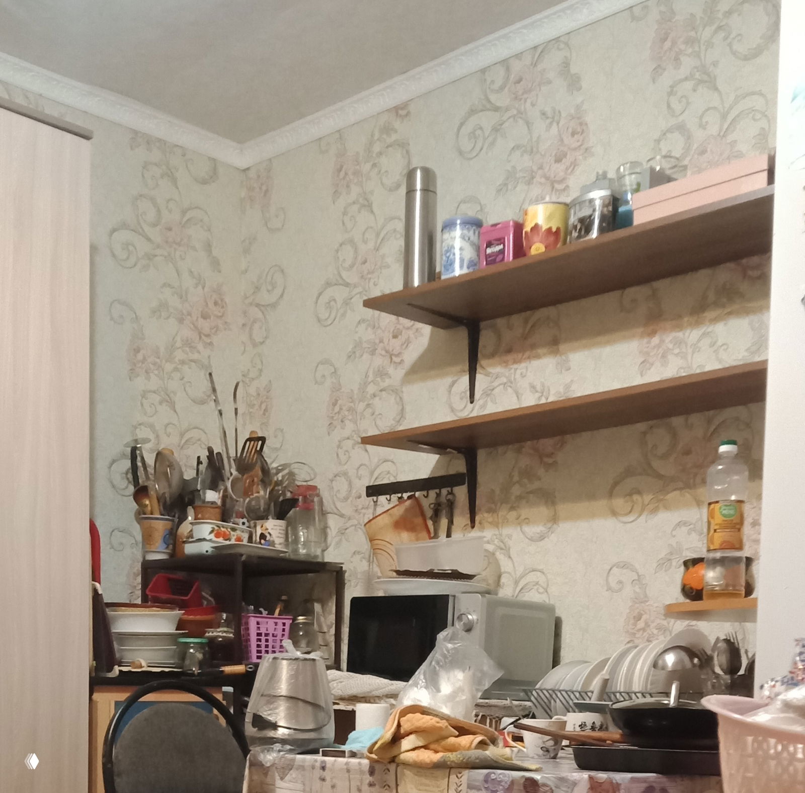 Кухня без закрытых шкафов: открытые полки с банками и баночками, микроволновка, стол с посудой, кухонными аксессуарами и разбросанными вещами в рабочем пространстве.