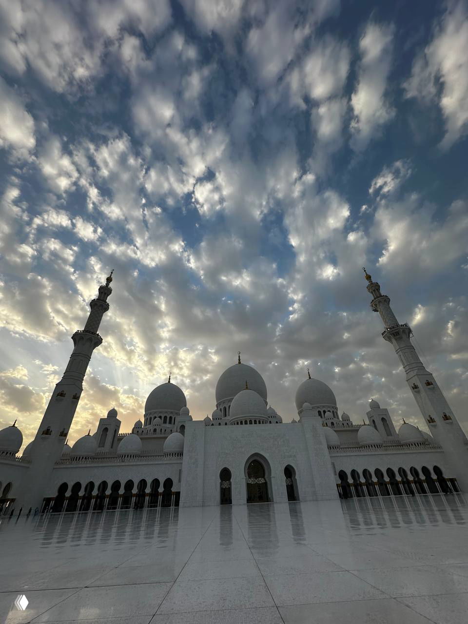 Фасад и купола большой белой мечети на фоне драматичного неба с облаками и лучами света; панорамный вид площади перед строением.