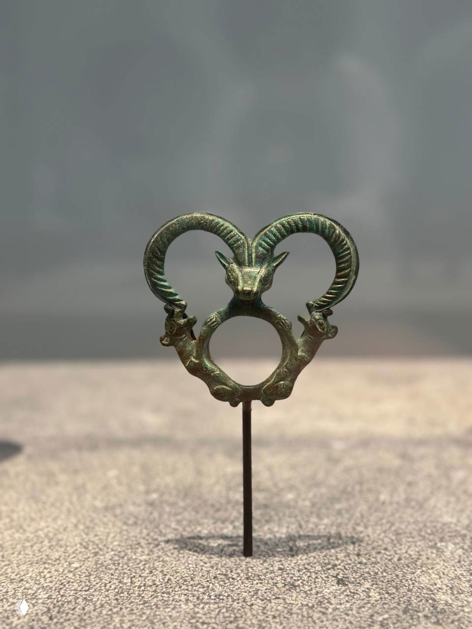 Музейный экспонат — небольшая бронзовая фигурка или украшение с рогами, установленная на подставке и снятая крупным планом на нейтральном фоне.