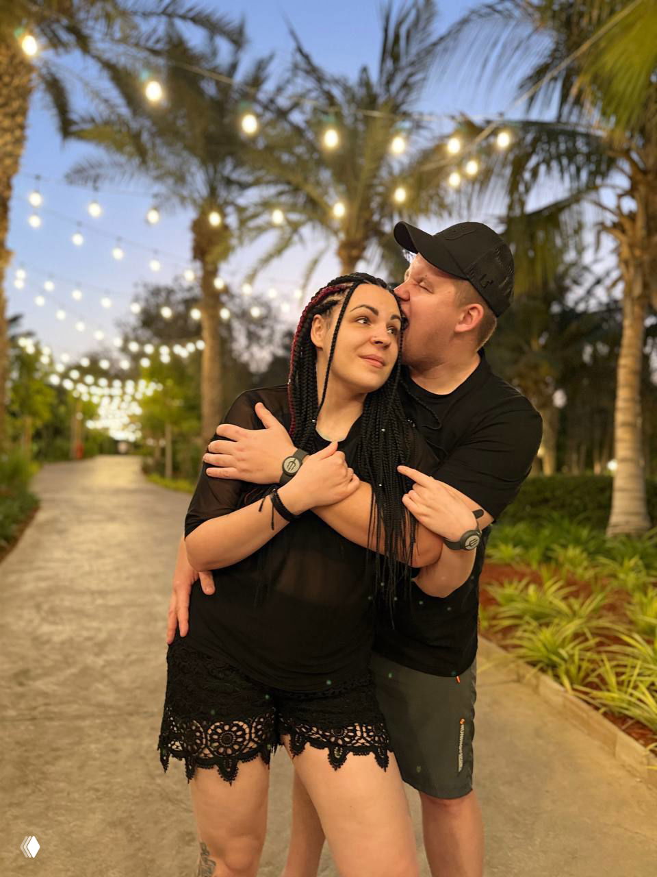 Пара обнимается на дорожке парка с гирляндами огней и пальмами; тёплая вечерняя атмосфера с размытой подсветкой и растительностью вокруг.