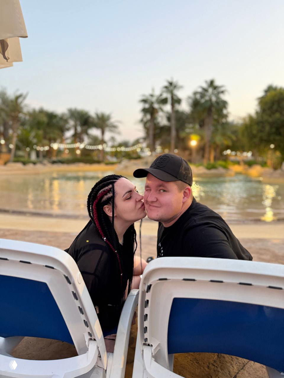 Пара сидит на шезлонгах у искусственной лагуны или бассейна в аквапарке/отеле: девушка целует мужчину, пальмы и вода на заднем плане в сумерках.