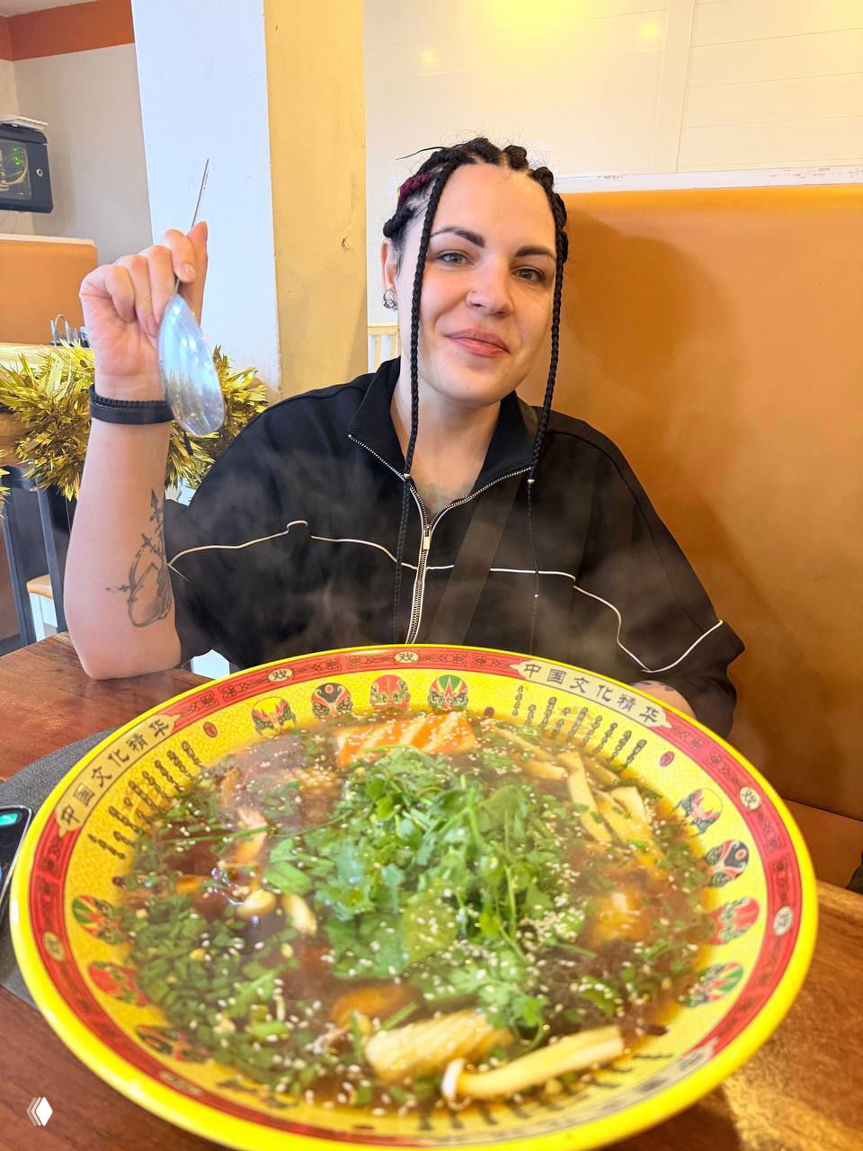 Автор за столом в ресторане с большой тарелкой супа или лапши на переднем плане; из блюда поднимается пар, видно интерьер заведения и улыбку автора.