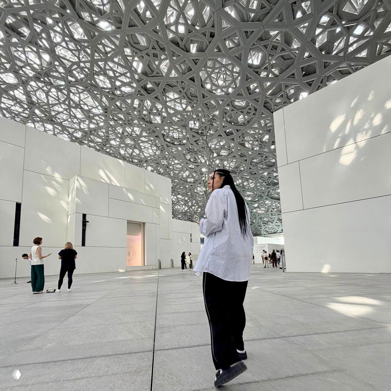 Женщина в полосатой рубашке идёт по просторному залу с орнаментным резным потолком; на заднем плане белые стены и прохожие.