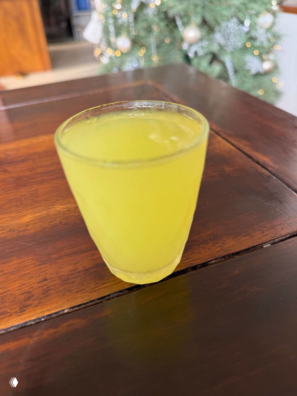 Небольшой рюмочный стакан с ярко-желтым лимончелло на деревянном столе; на заднем плане размытая рождественская елка и огни.