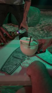Крупный план подачи: маргарита в брутальном бокале с перевёрнутой бутылкой пива, руки бармена и меню на столе.