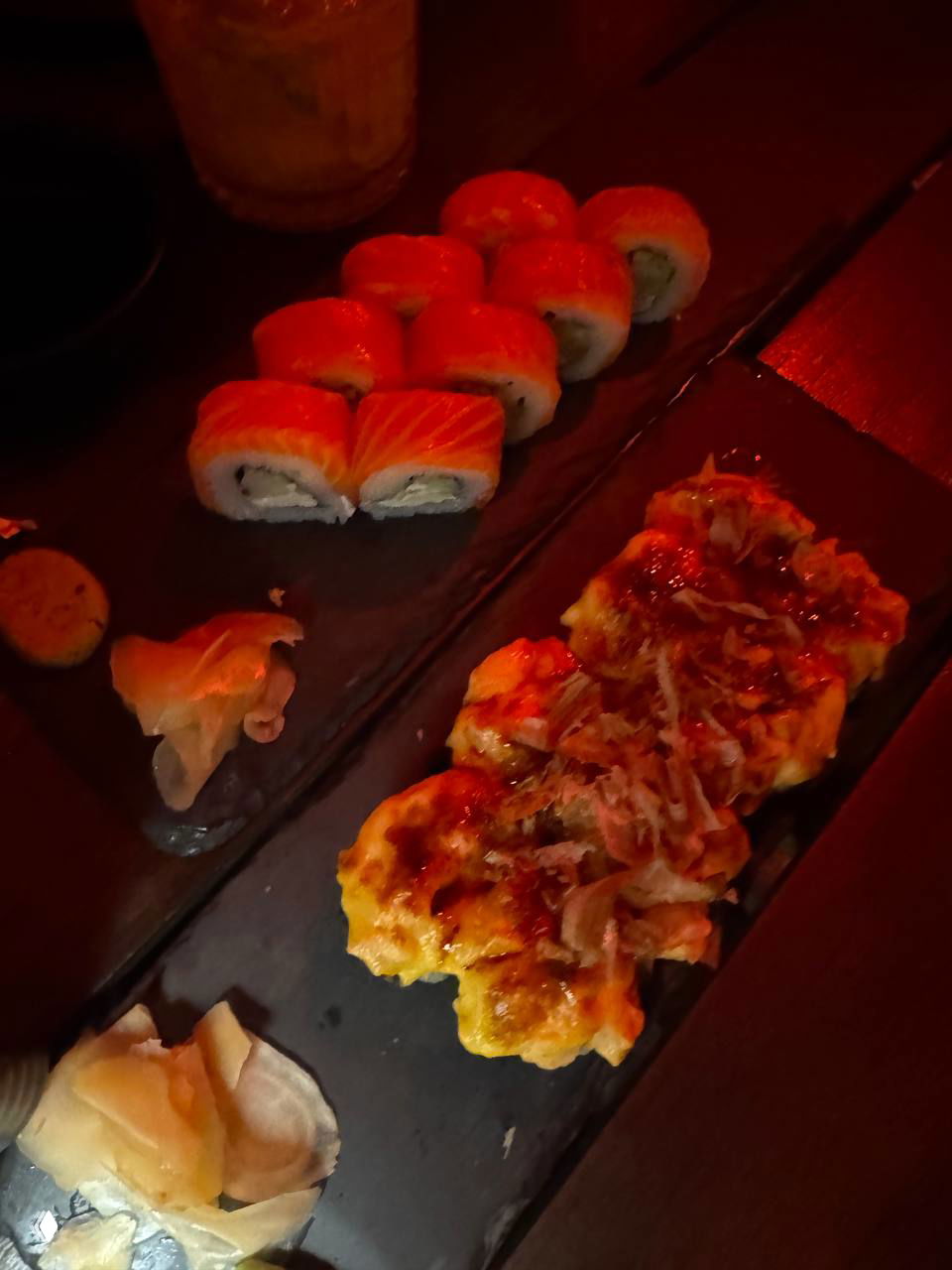 Чёрная сервировочная доска с роллами «Филадельфия» и запечёнными роллами, соусом и кусочками имбиря, снято при приглушённом красном освещении ресторана.