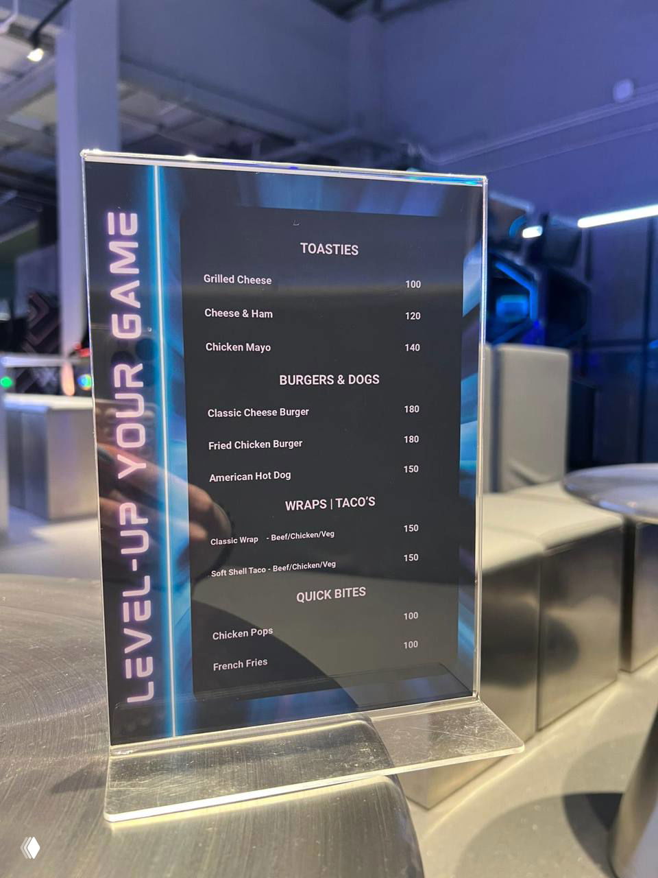 Ближайший план на настольную табличку-меню зоны игр: разделы Toasties, Burgers & Dogs, Wraps и Quick Bites с ценами в бат и отражением вокруг.