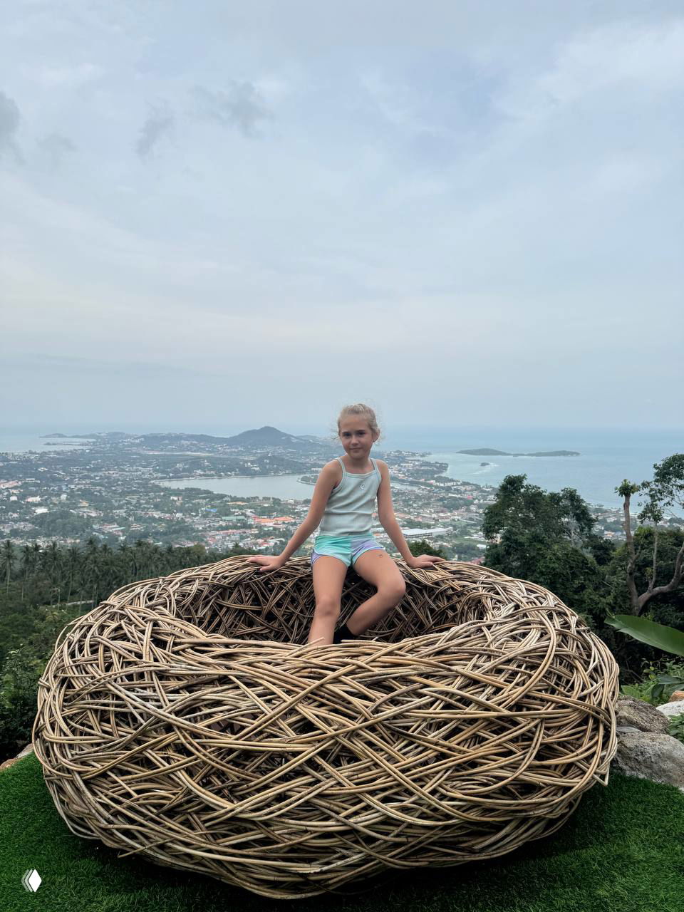 Большое плетёное гнездо-Фотозона, в котором сидит ребёнок на фоне панорамного вида на Самуи и прибрежную линию.