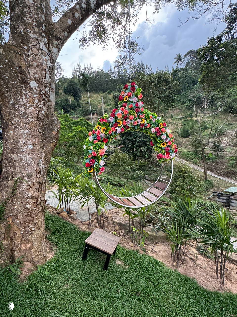 Подвесное кресло-круг в цветочных гирляндах, висящее у деревьев над зелёным склоном с тропинками.
