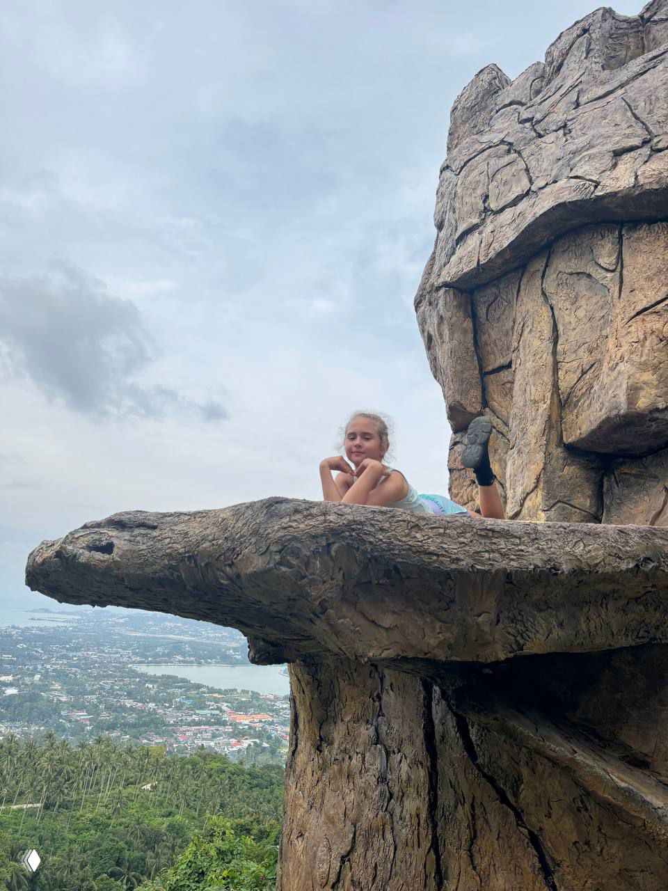 Ребёнок лежит на выступе искусственной скалы над объёмным панорамным видом — опасный ракурс для снимка на краю обрыва.
