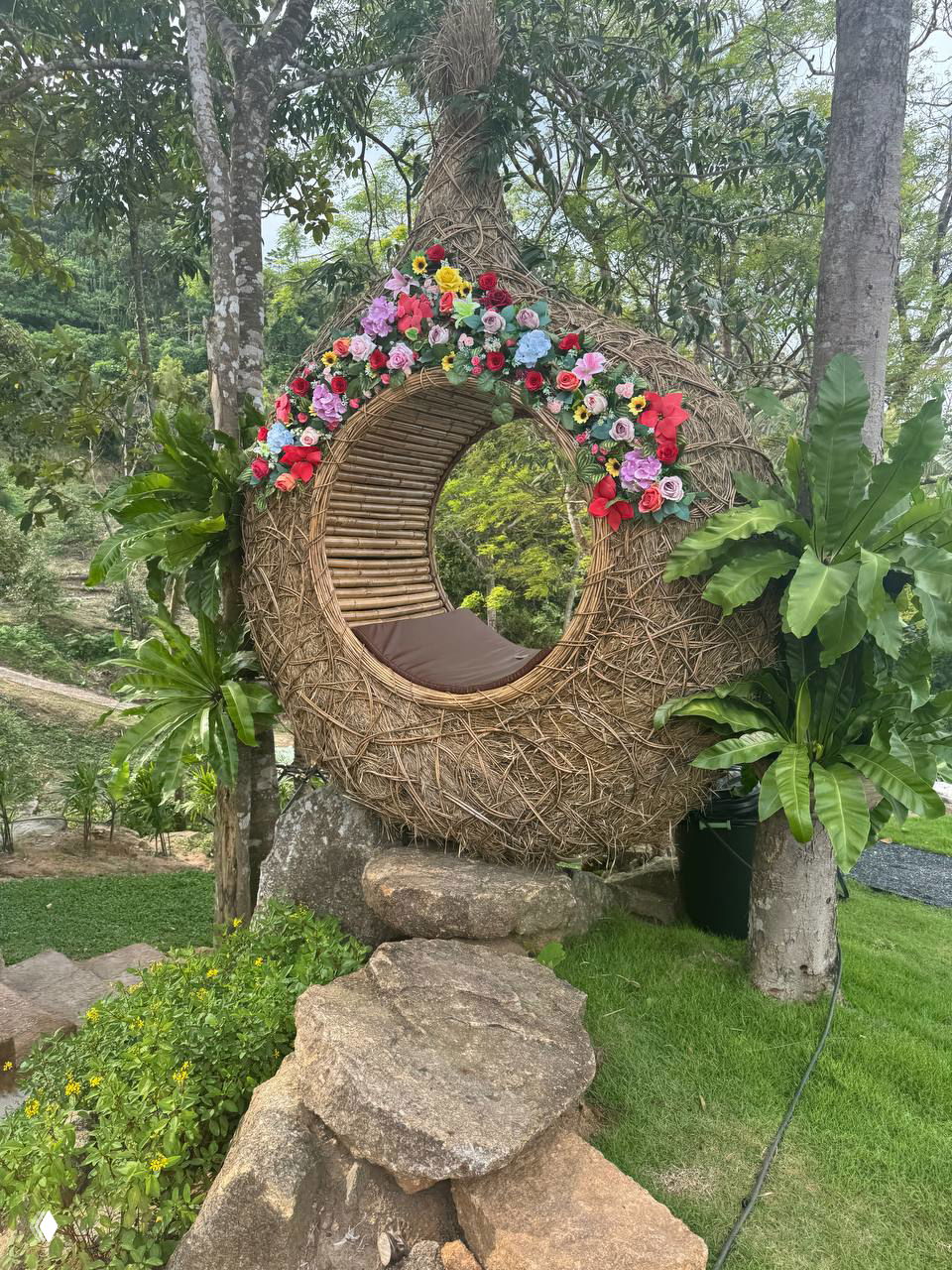 Круглая плетёная кабина-подушка, украшенная цветами, размещённая на камнях и траве для уединённых фото.