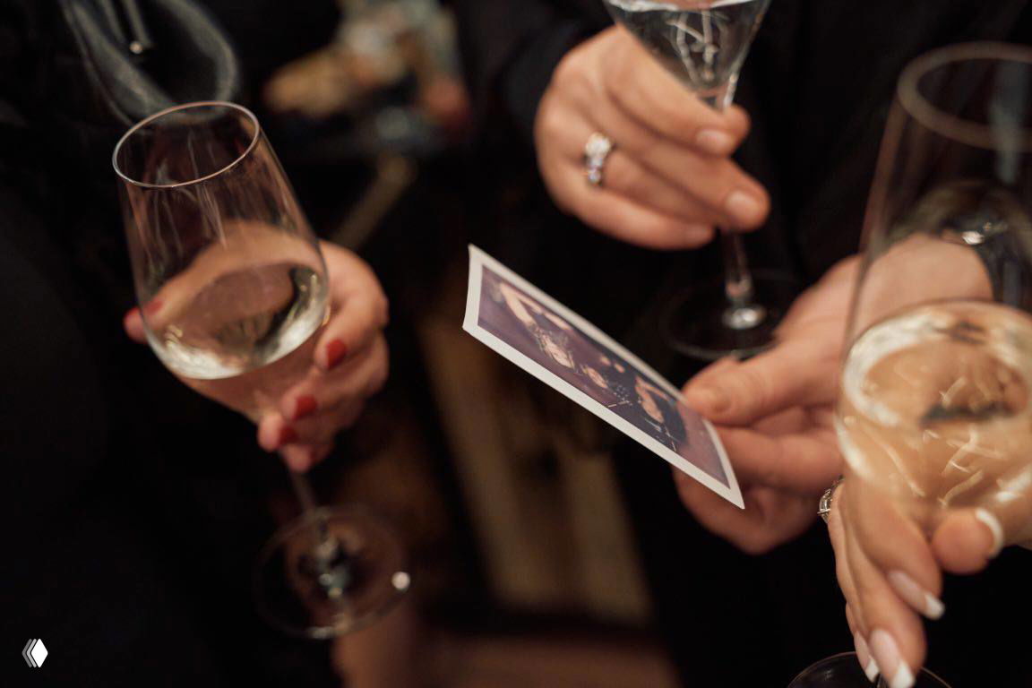 Гости рассматривают только что распечатанную моментальную фотографию, держат бокалы с вином — живой кадр вечернего события.