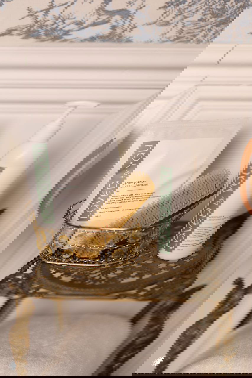 Продуктовая композиция Loved by You: пакет скраба, бутылка молочка и щётка на декоративном столике в ванной; эстетика домашнего ухода.