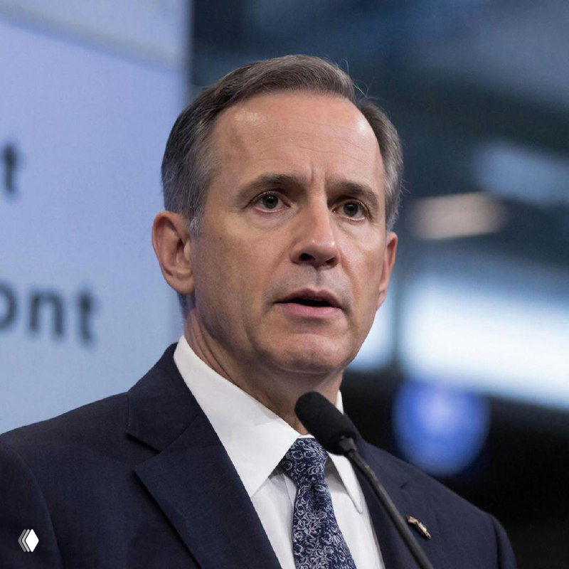 Портрет мужчины в деловом костюме и галстуке у микрофона на фоне конференц-зала; деловая атмосфера и нейтральный размытый фон, иллюстрация пресс‑конференции.