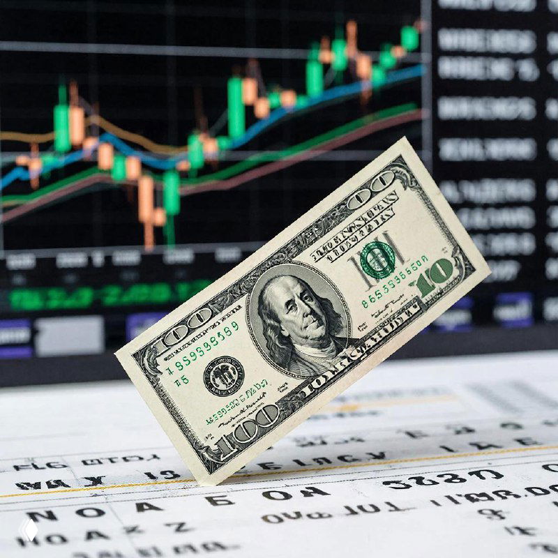 Купюра доллара на переднем плане на фоне свечного графика и финансовых таблиц — иллюстрация давления на валютный рынок и возможной волатильности.