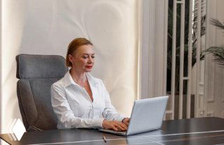 Женщина средних лет в белой рубашке сидит за столом с ноутбуком в светлом офисе; деловая поза и рабочая атмосфера, интерьер кабнета виден в кадре.