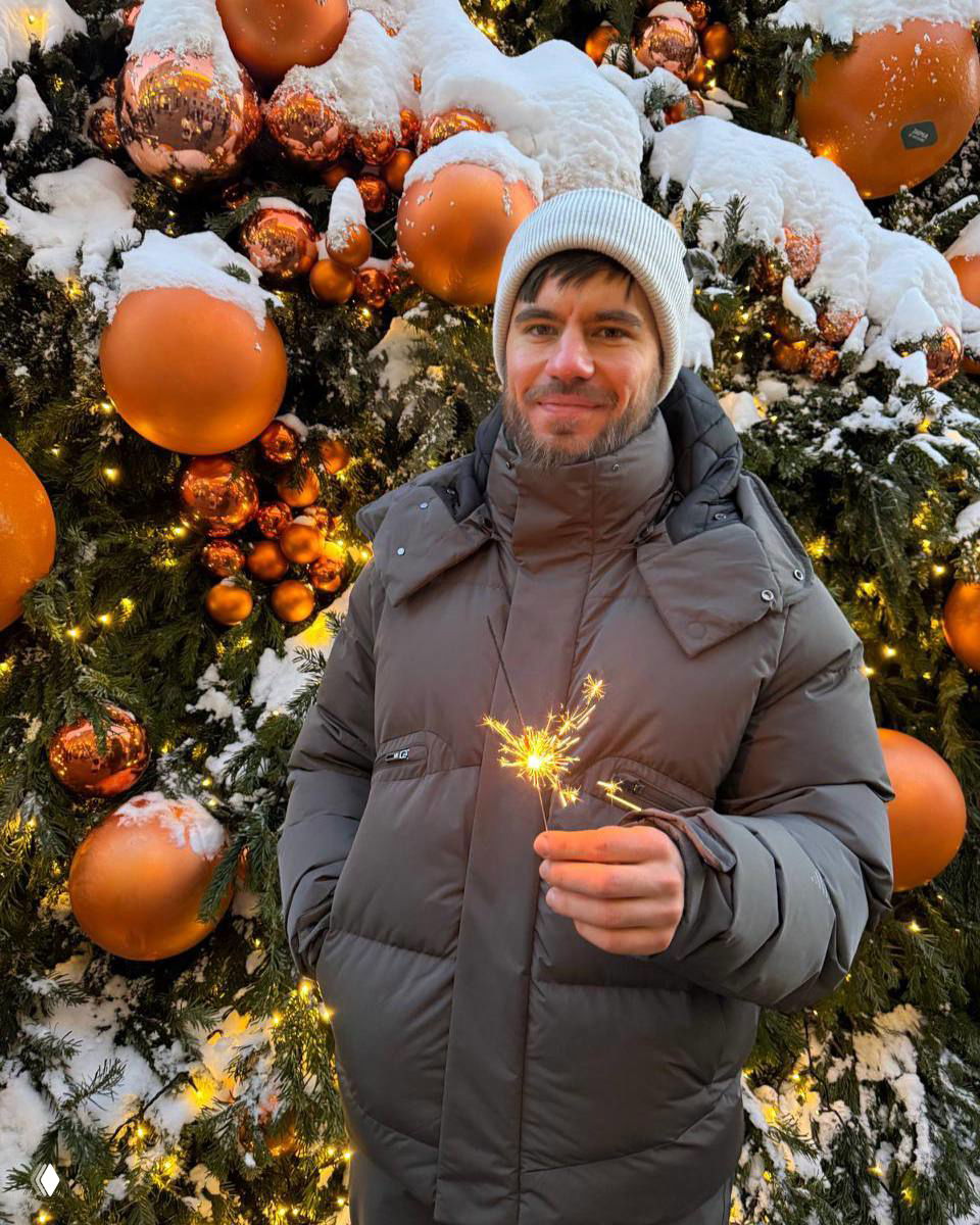 Человек в зимней куртке и шапке держит бенгальский огонь на фоне украшенной рождественской ёлки с большими оранжевыми шарами и снегом.