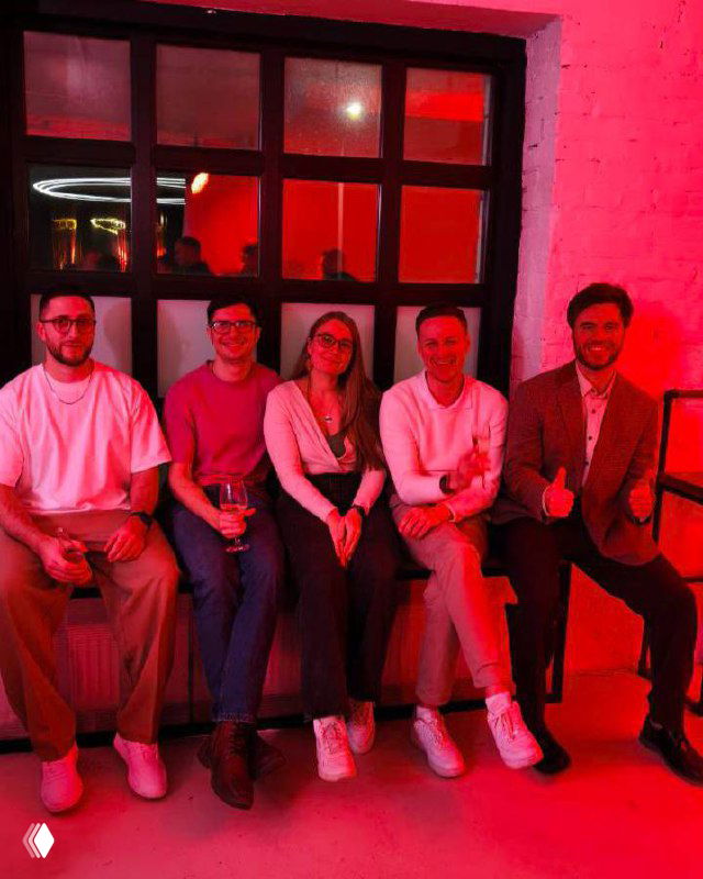 Пятеро коллег сидят на скамье в комнате с красной подсветкой; неформальная встреча, обсуждение проектов, обмен контактами.