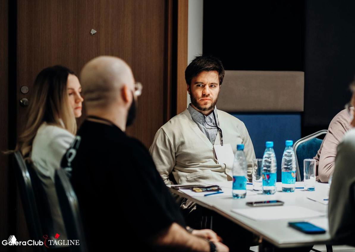 Участники деловой встречи сидят за столом, обсуждают маркетинговый кейс; на столе блокнот, телефоны и несколько бутылок воды — фото с конференции.