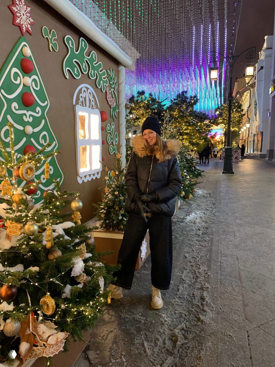 Женщина в пуховике стоит у домика с пряничным декором и светящимися гирляндами под переливающейся иллюминацией на улицах города.