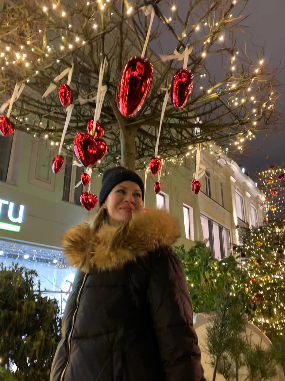 Гирлянды и сердечки в ветвях дерева над прогулочной аллеей; автор смотрит вверх, вокруг — огни и праздничные украшения ночного города.