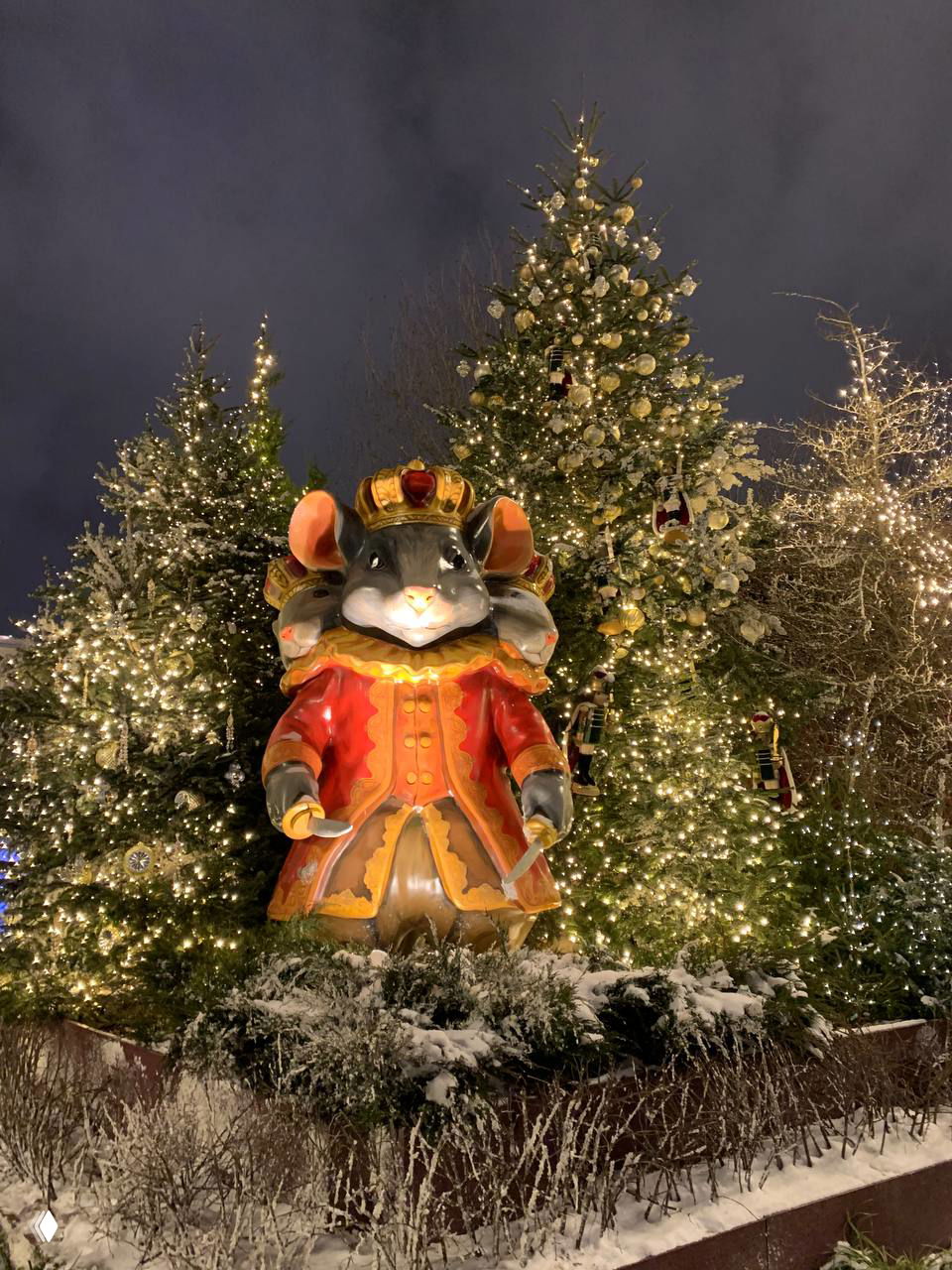 Ночное фото пешеходной улицы под потолком светящихся гирлянд и украшений, внизу — ограждение с нитями лампочек и праздничные ели.