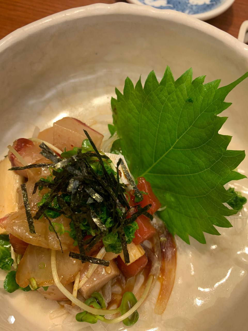 Небольшая порция маринованной сырой рыбы с листом шисо и мелко нарезанными ингредиентами на белой тарелке — закуска в локальном заведении.