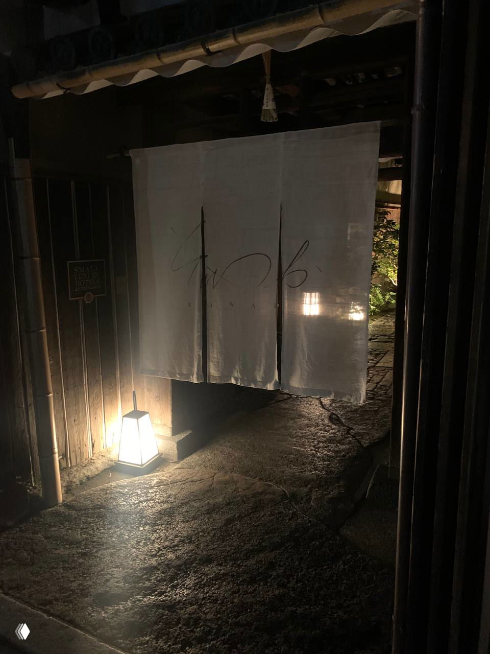 Белая тканевая занавесь норэн на входе в скромное заведение, освещённая мягким фонарём в тёмное время суток.