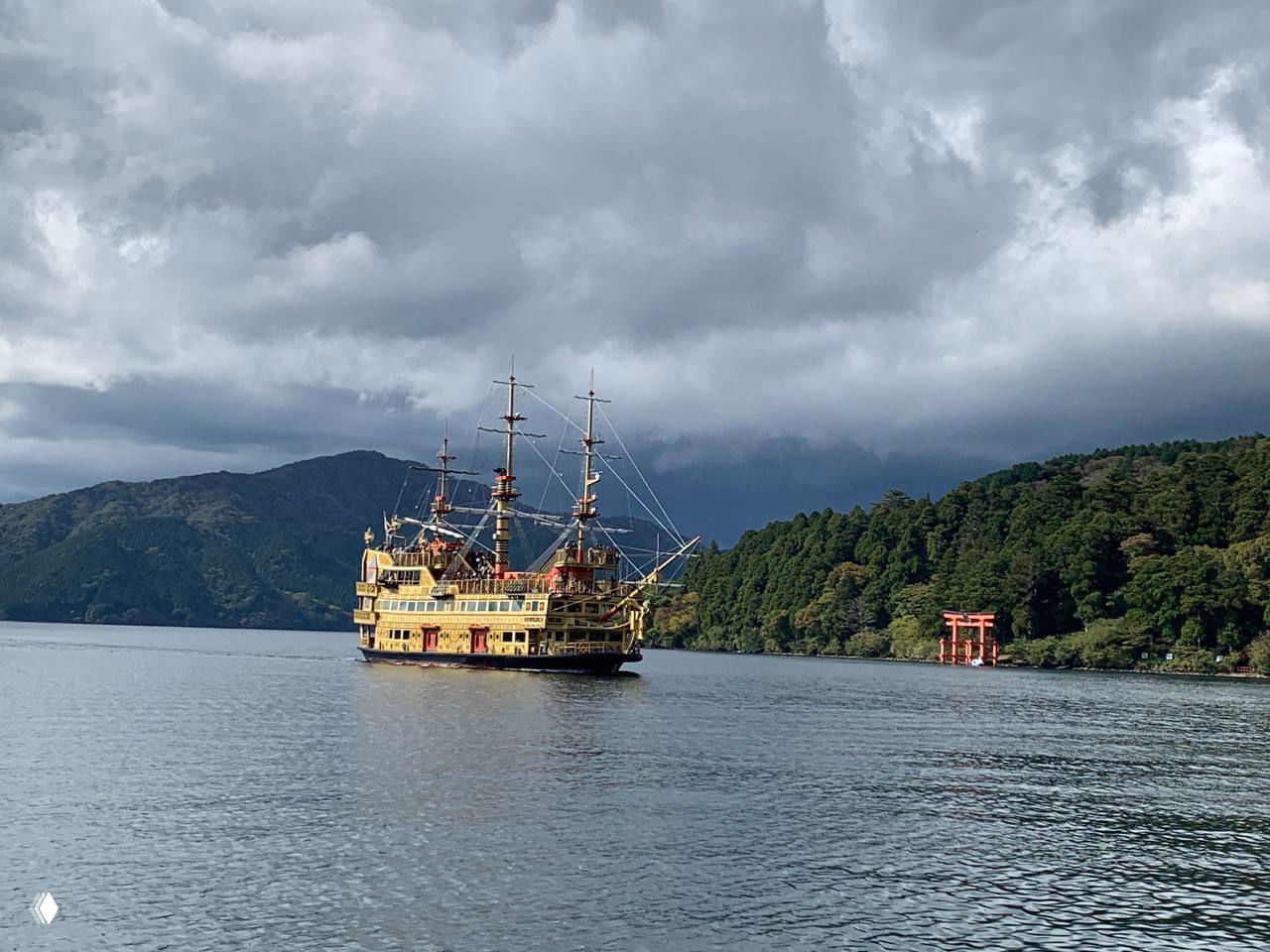 Парусник-экскурсионное судно на озере Аси: декоративный деревянный корабль плывёт по зеркальной поверхности озера, на горизонте лес и тории.