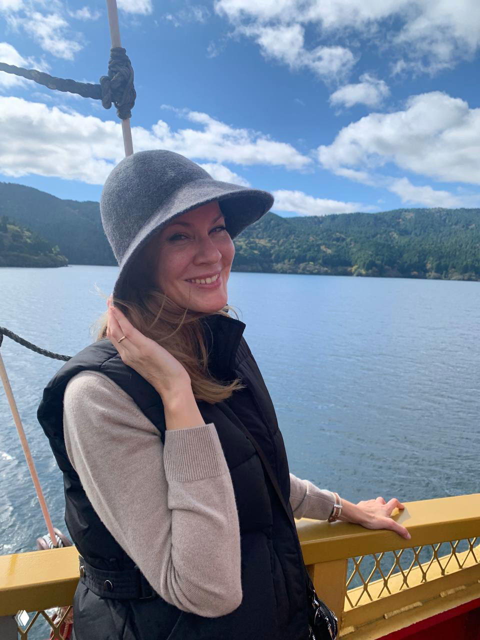 Портрет улыбающейся женщины на фоне озера в Хаконэ: туристка в шляпе стоит на палубе, позирует на фоне водной глади и лесистых склонов.