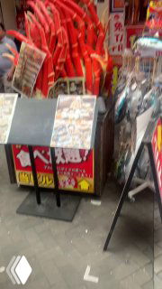 Уличная торговая зона с рекламными стендами, товарами и неоновыми элементами, создающая плотную среду внимания и стремление пройти внутрь заведений.