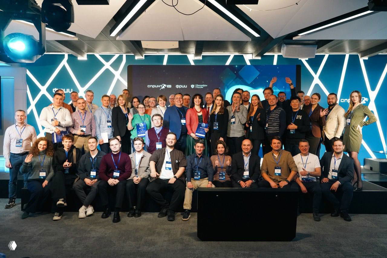 Групповое фото участников акселератора на сцене итогового мероприятия: команды и трекеры на фоне брендового экрана и световой декорации.