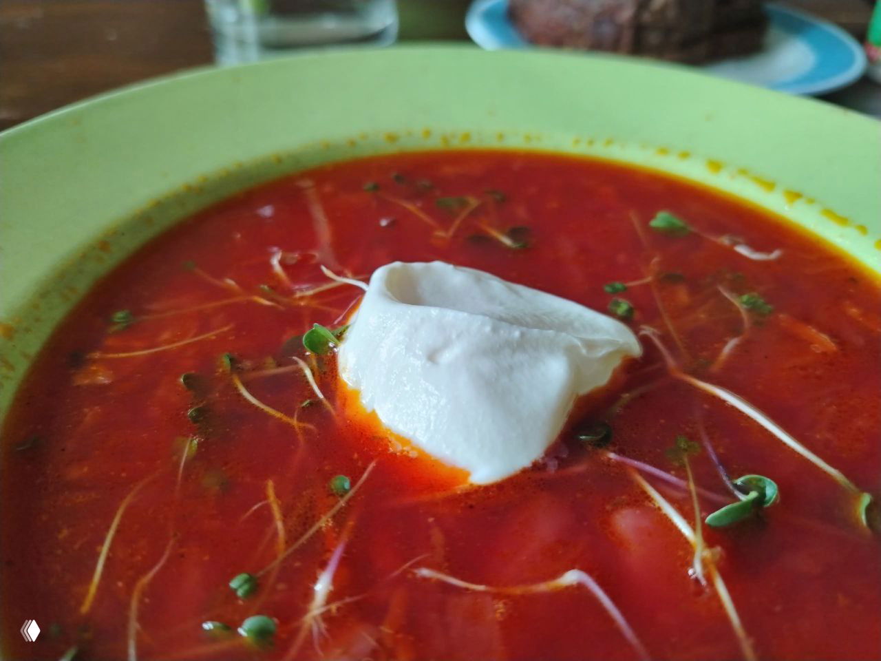 Крупный план тарелки с насыщенно-красным супом (борщ или свекольный суп) с ложкой сметаны и зеленью, домашняя еда в уютной обстановке.