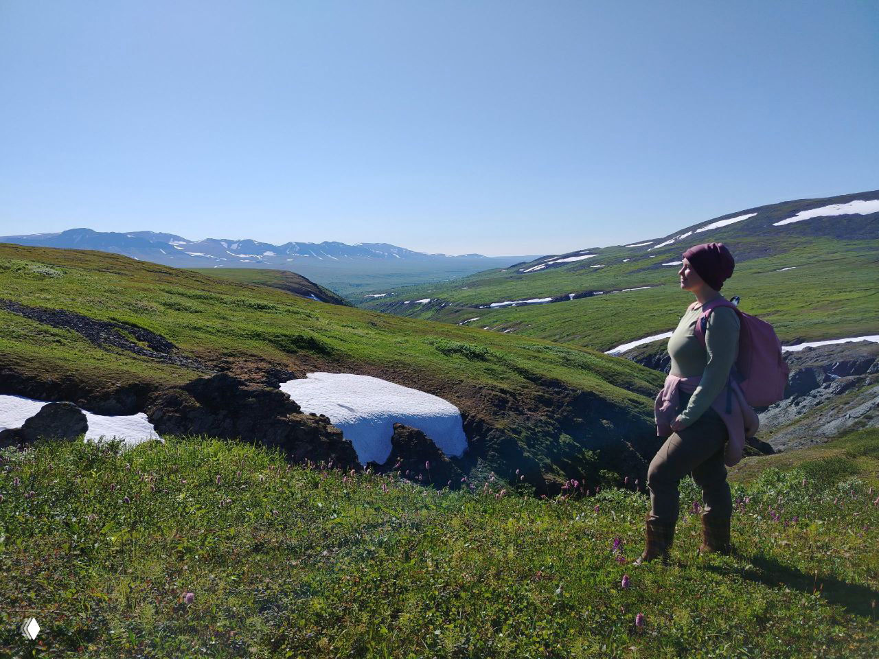 Пейзаж Полярного Урала: горные луга с остатками снега, дальние хребты и женщина в походной одежде с рюкзаком смотрит вдаль на долину.