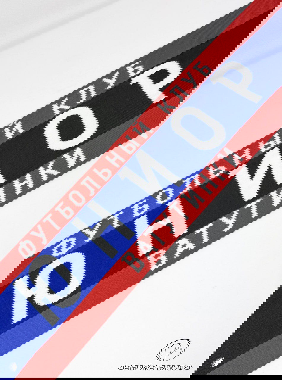 Длинный план шарфа синего цвета с красными краями и белыми буквами «ЮНИОР» и указанием Ватутинки на белом фоне, акцент на дизайне надписи.