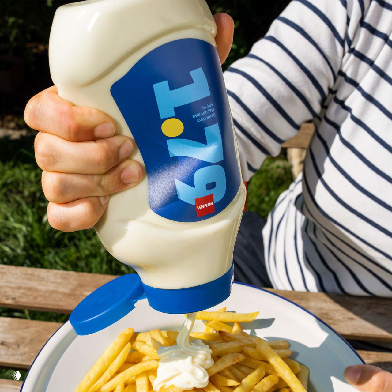 Рука держит бутылку майонеза с яркой этикеткой и крупной цифрой цены; соус выдавливается на тарелку картофеля фри на открытом воздухе.