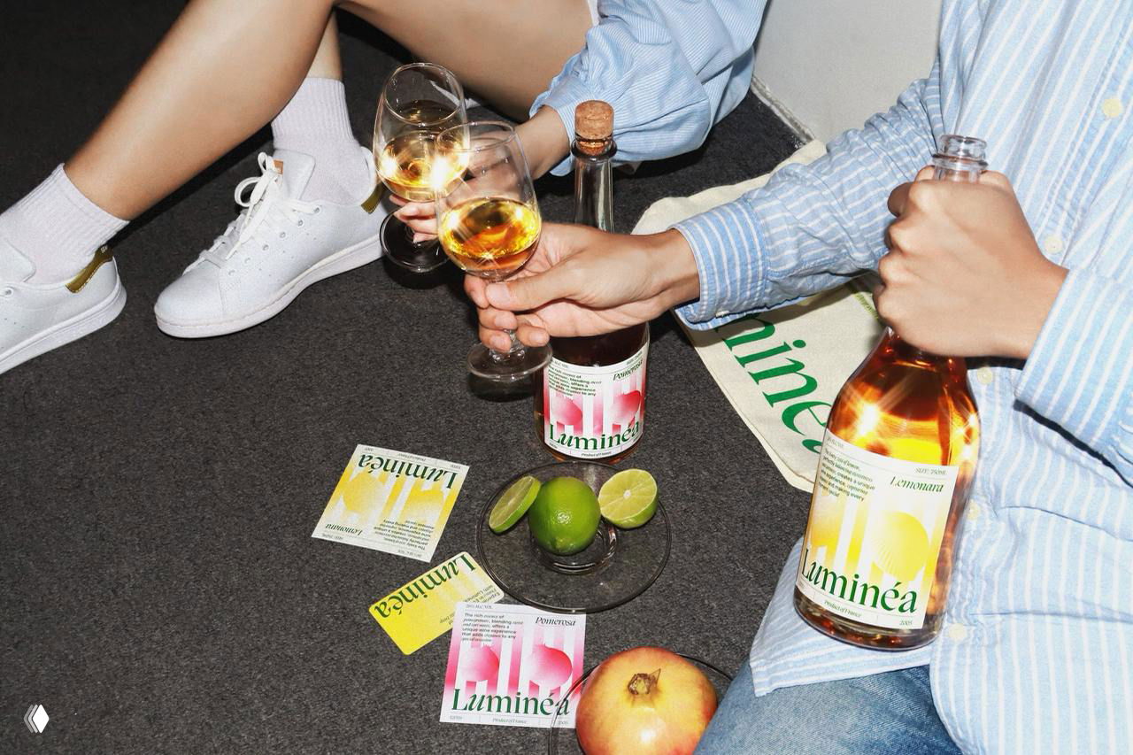 Фотография: бутылка Luminea и бокалы в руках двух людей на полу, рядом этикетки, лайм и яблоко — светлая молодёжная стилистика с градиентами.