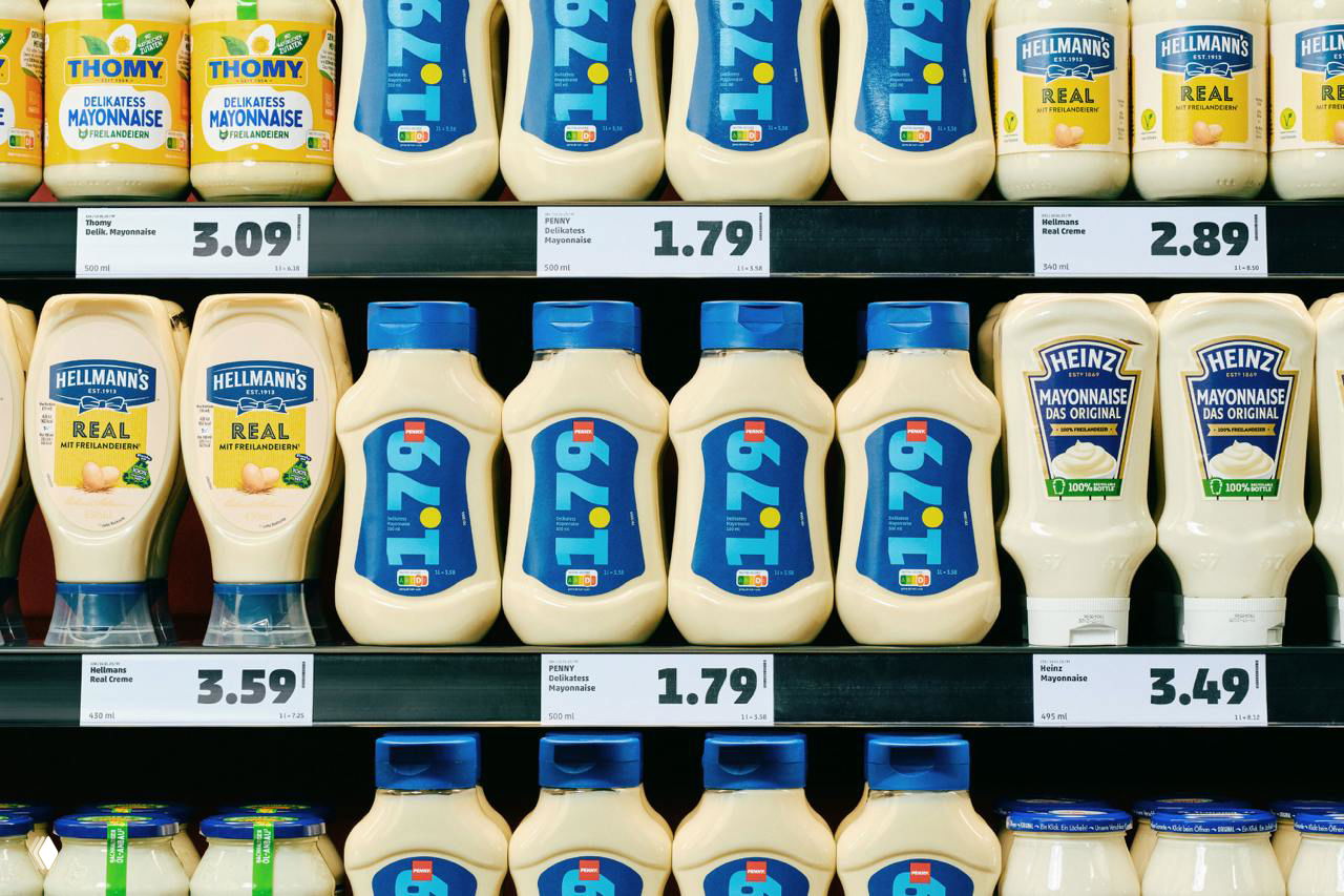 Ряды бутылок майонеза на торговой полке: бренд Penny с синими этикетками и крупными ценниками на полке под продуктами.