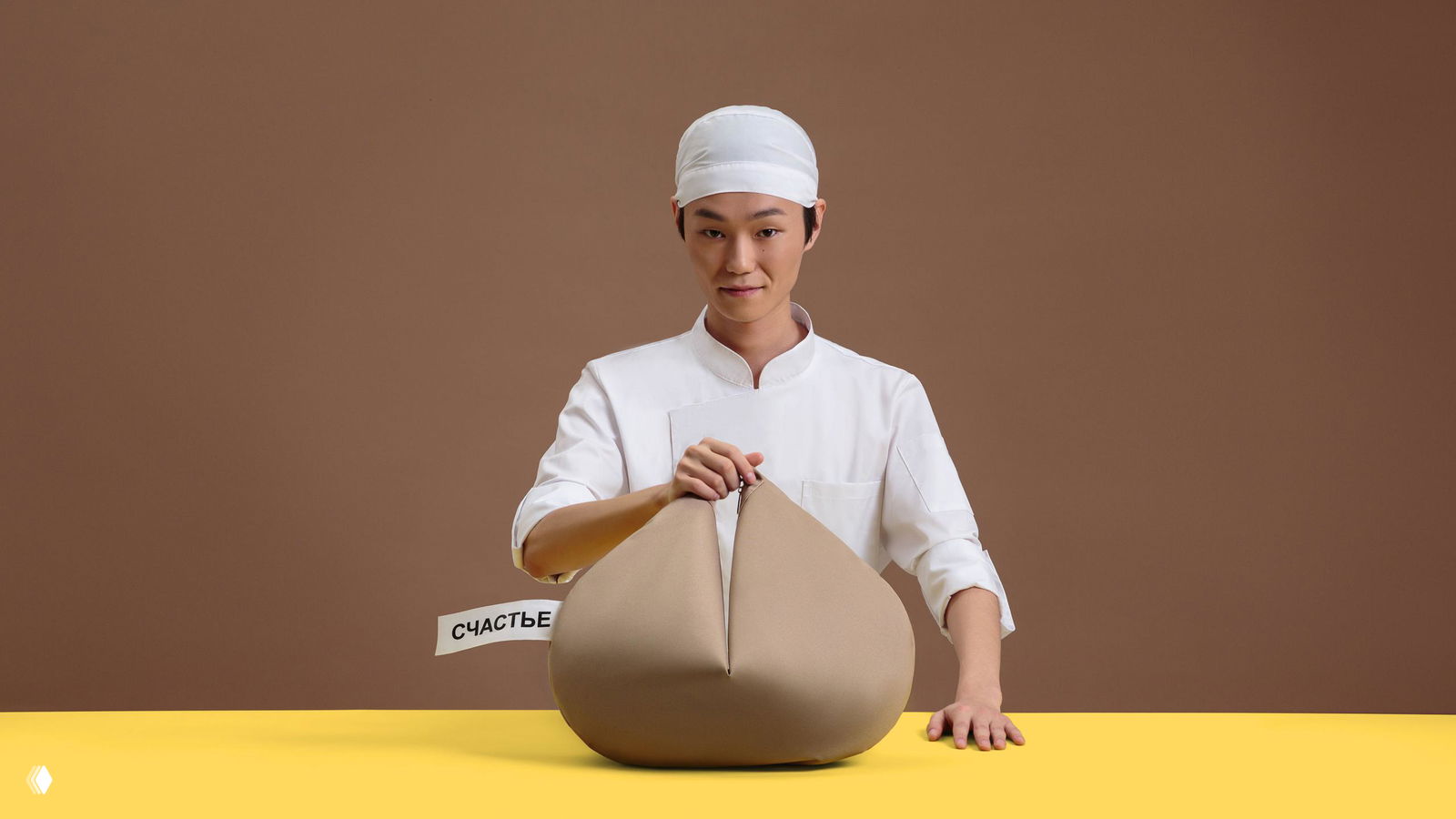 Кадр с моделью за столом: повар или стилизованная персона держит круглую сумку‑печенье на жёлтом столе; видно белый хвостик с надписью «СЧАСТЬЕ» и бирку бренда.