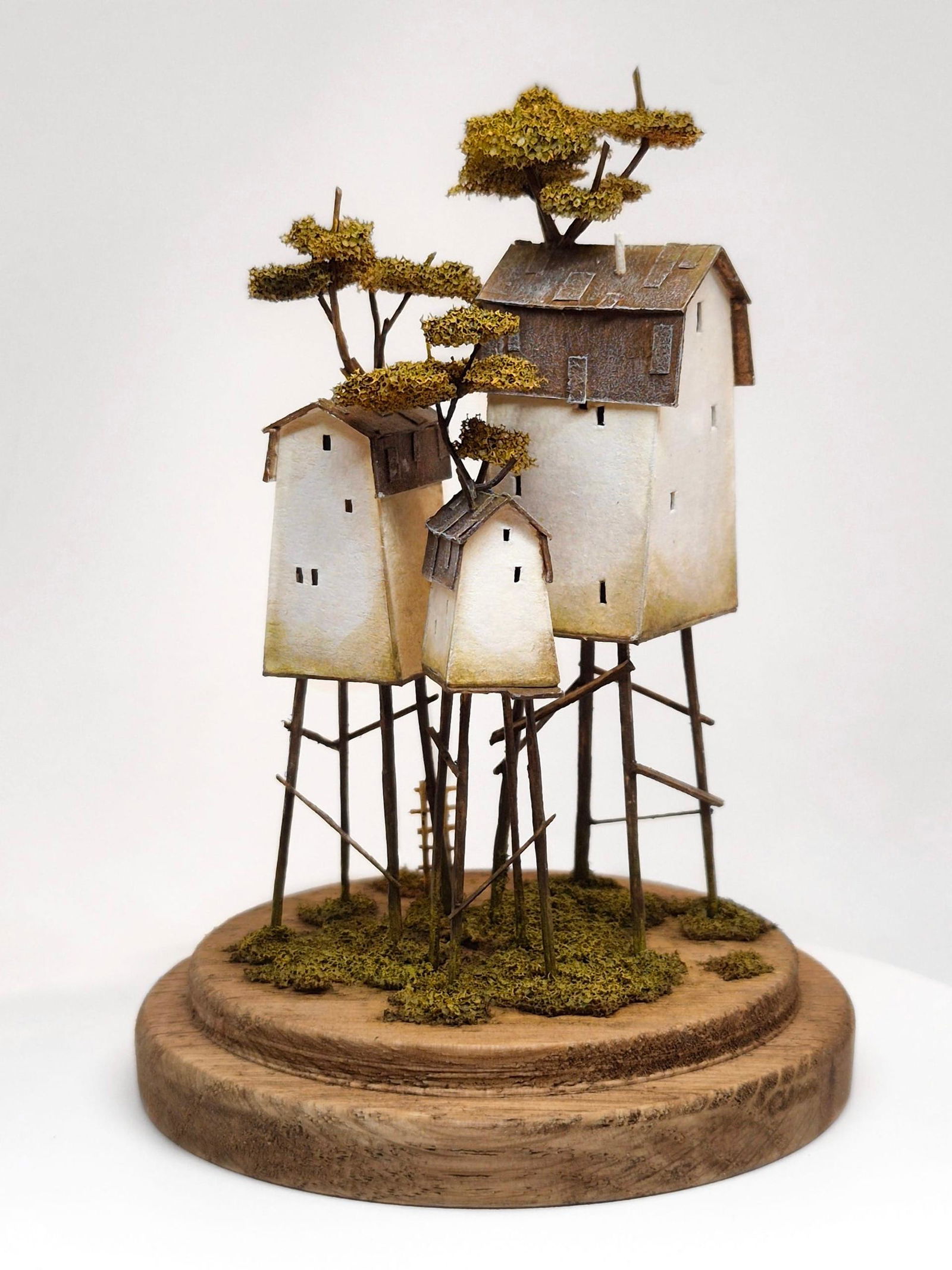 Небольшая группа миниатюрных домиков на деревянной основе с опорами‑жердями и аккуратно размещенным мхом — отточенная модельная работа художника.