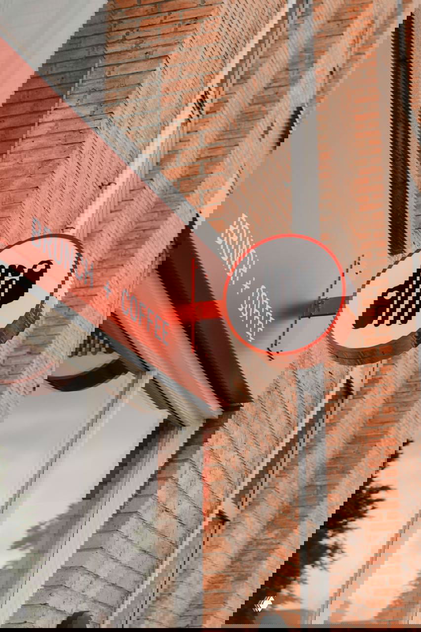 Круглый наружный знак кафе с логотипом Drip на кирпичной стене и красной вывеской «BRUNCH + COFFEE», спокойная уличная сцена в фирменных цветах.