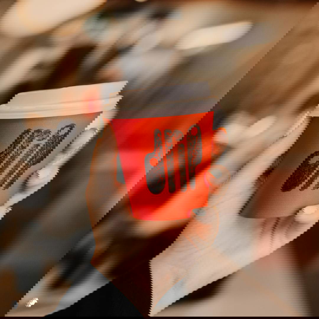 Рука держит красный бумажный стакан с фактурным оттиском логотипа Drip; кадр подчёркивает «стекающую» типографику и тёплую интерьерную гамму.