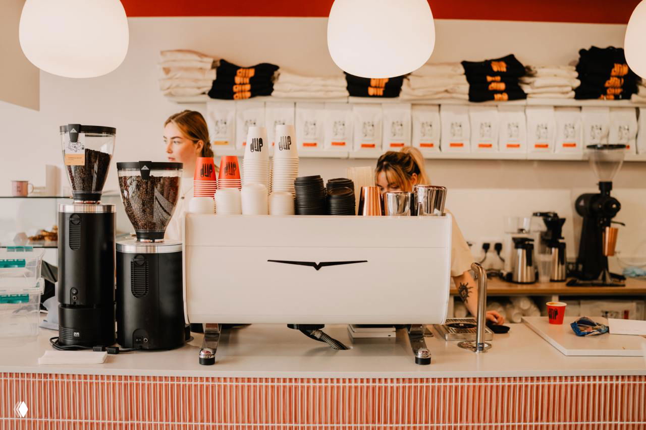 Интерьер кофейни: бариста за стойкой, кофемашина, стопки брендированных стаканов Drip и аккуратная выкладка товаров в фирменных цветах и стиле.