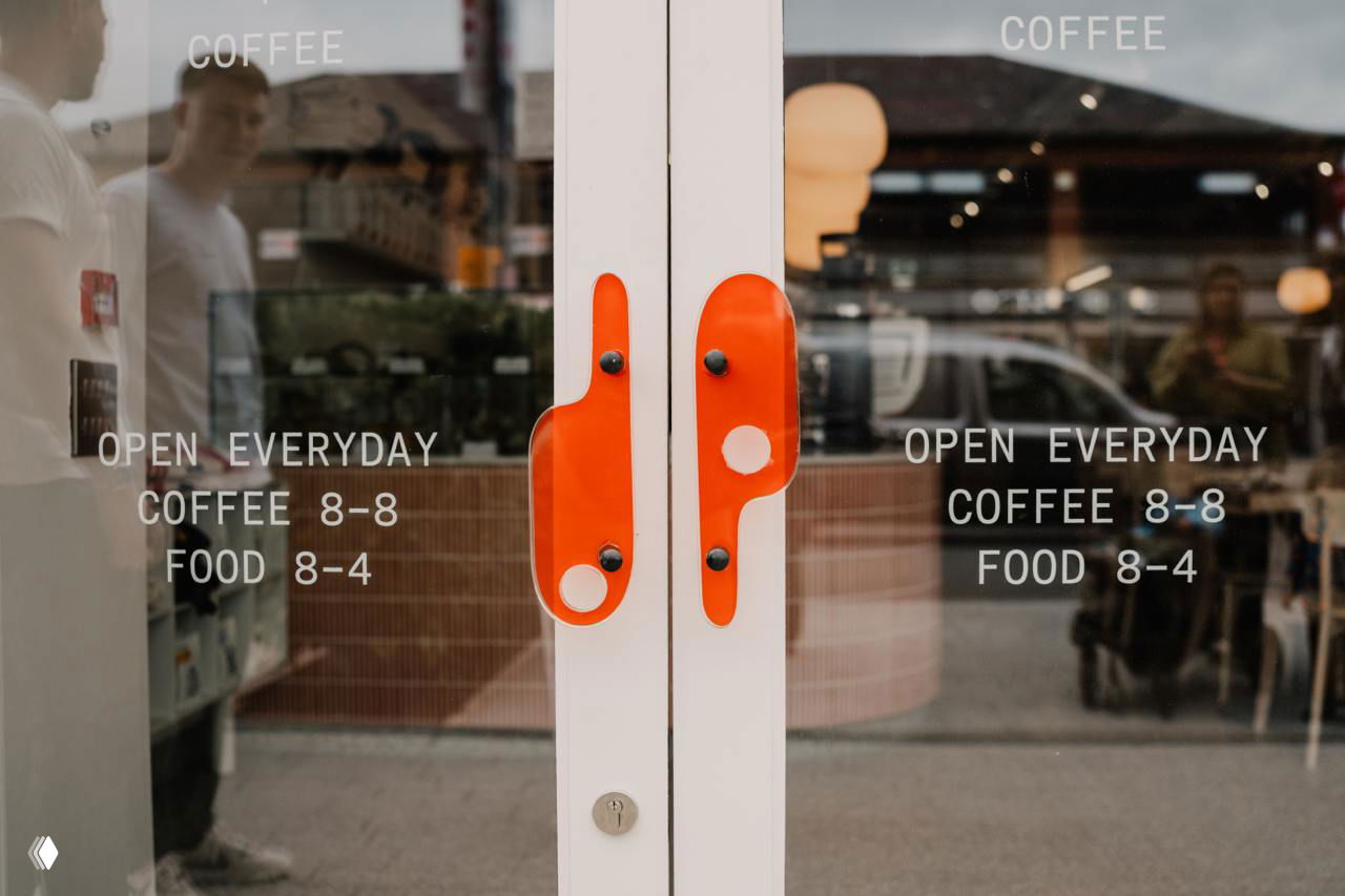 Стеклянная входная дверь с ярко-красными ручками и надписями «OPEN EVERYDAY COFFEE 8-8 FOOD 8-4», видны отражения улицы и стильное оформление входа.